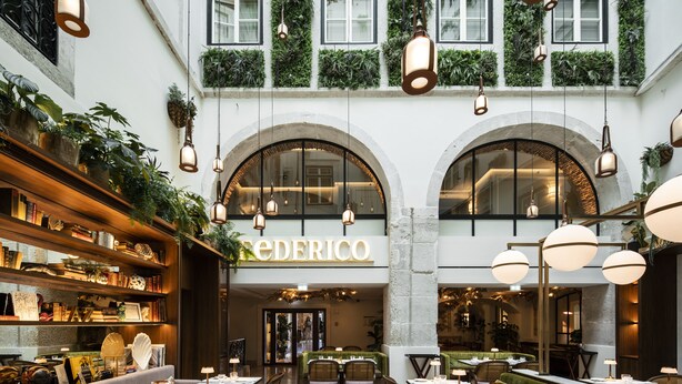 Ein Restaurant mit einem zentralen, offenen Essbereich, mehreren Tischen und Stühlen, umgeben von Bücherregalen und Grünpflanzen.