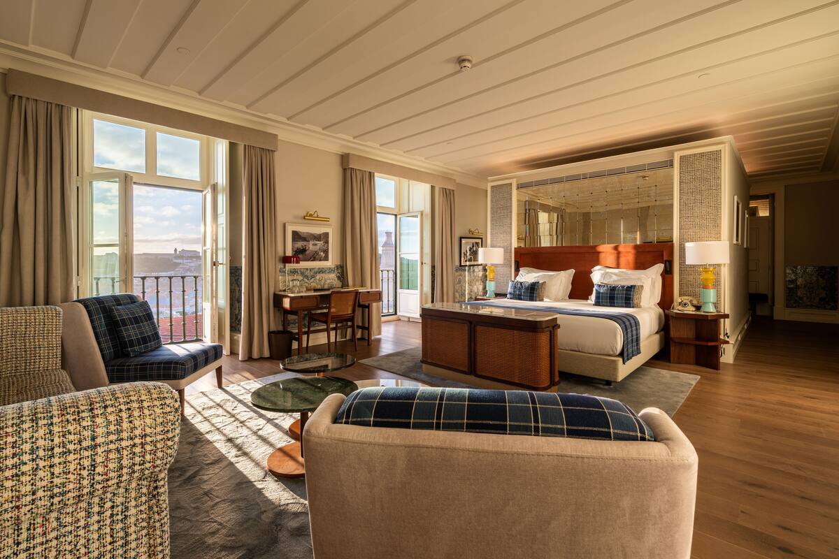 Ett rymligt hotellrum med en stor säng, en sittgrupp med soffa och fåtölj, en matplats med bord och stolar samt utsikt över staden genom stora fönster.