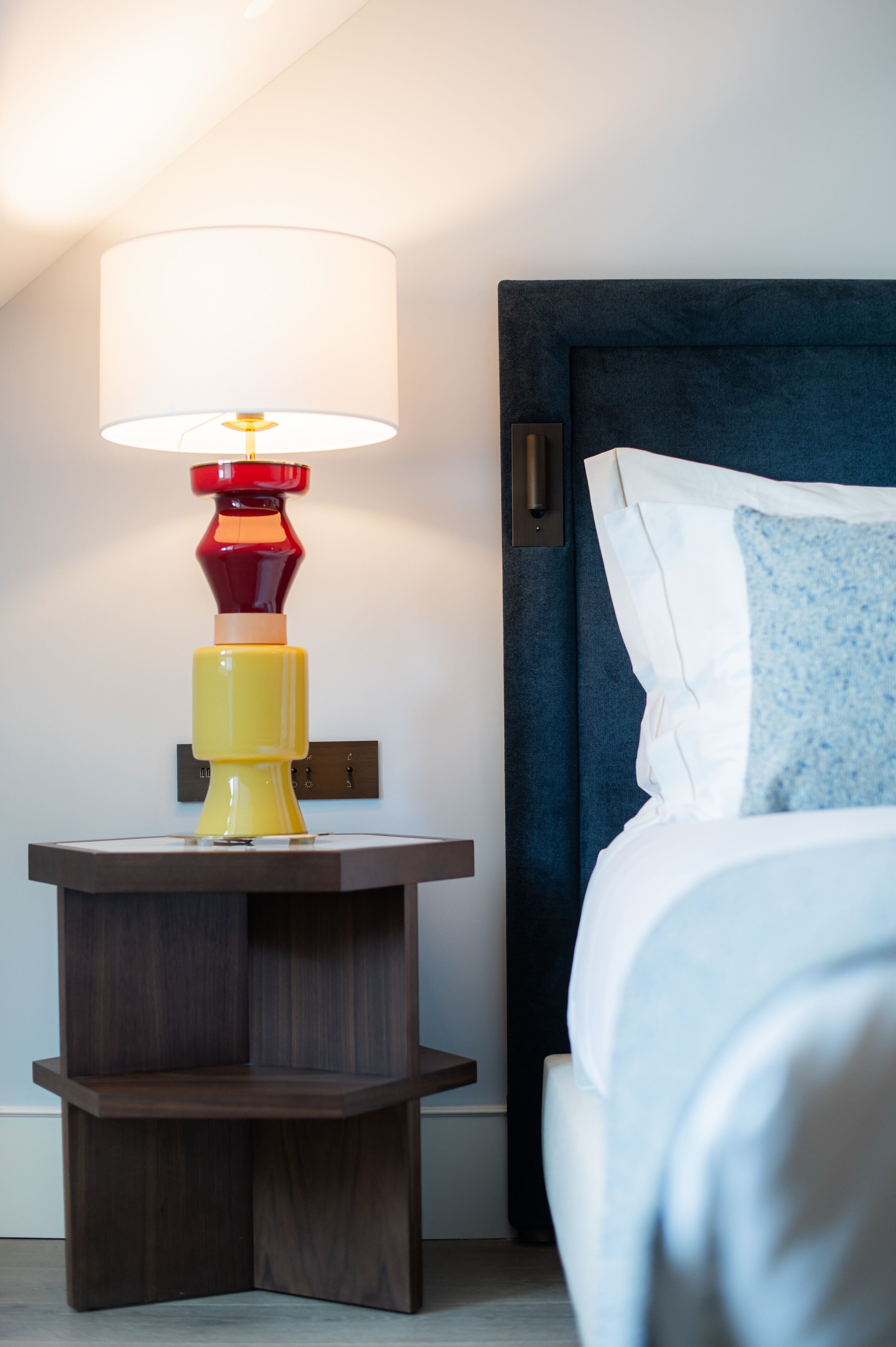 A neatly made bed with a blue pillow and a white pillow, a bedside table with a lamp, and a dark blue headboard.