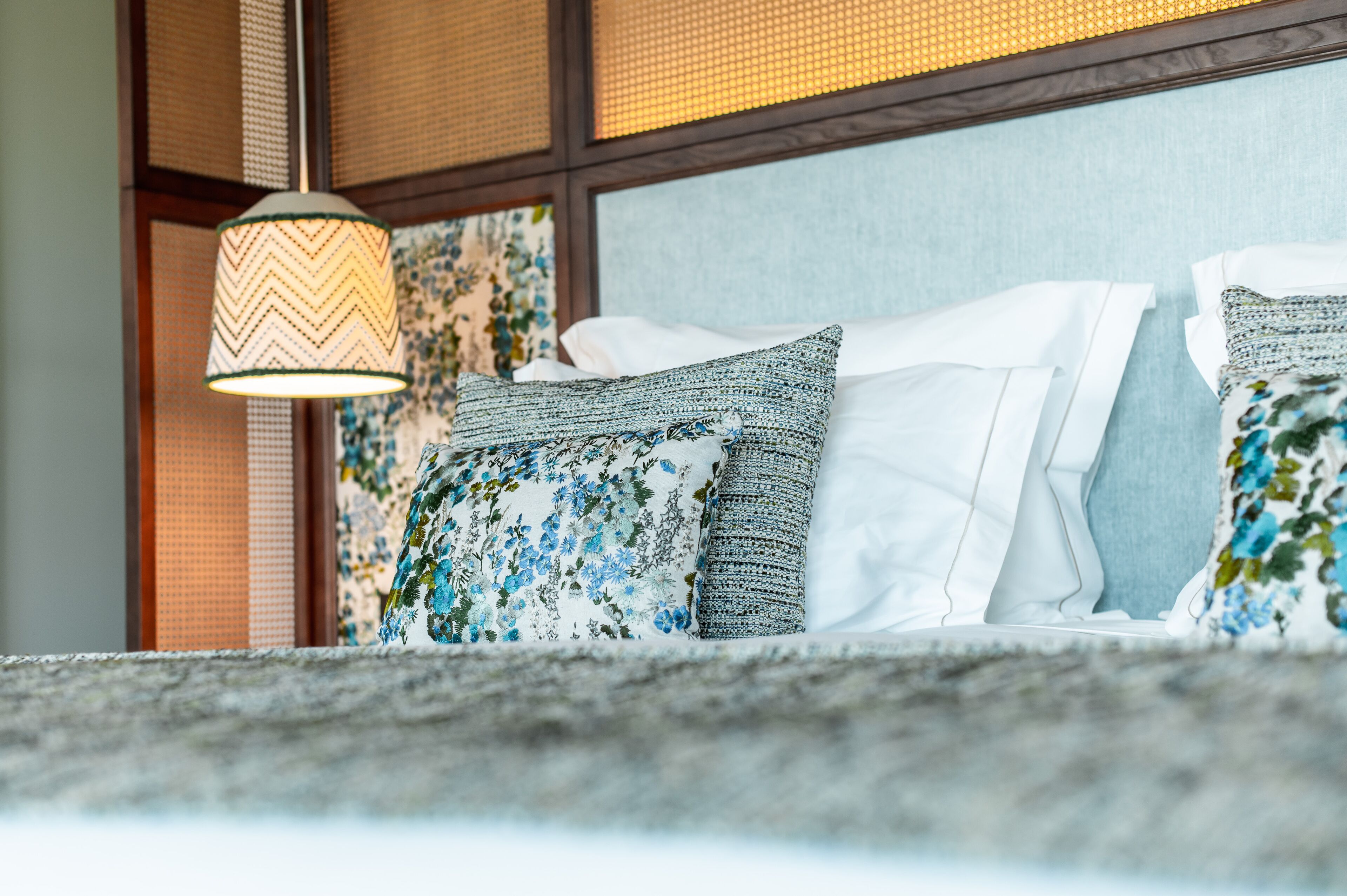 Una cama con almohadas estampadas y una lámpara colgante en una habitación con pared texturizada y una ventana con persianas.