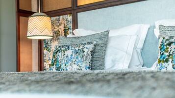 Una cama con almohadas estampadas y una lámpara colgante en una habitación con pared texturizada y una ventana con persianas.