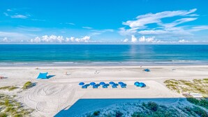 Ein Strand mit einer Reihe blauer Sonnenschirme und Liegestühlen, ein sandiger Bereich mit Radspuren und einem wolkenlosen, blauen Himmel.