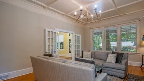 A living room with a large sectional sofa, a glass coffee table, and a grey sofa with cushions. There is a chandelier hanging from the ceiling and large windows with white frames.