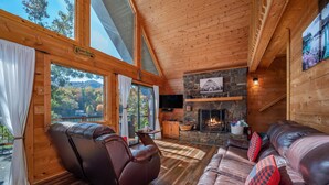 L’intérieur chaleureux d’un chalet avec une cheminée en pierre, des canapés en cuir et de grandes fenêtres offrant une vue panoramique.