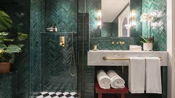 A bathroom with a herringbone tile pattern, a glass shower enclosure, a white sink with gold handles, and a red bench with towels.