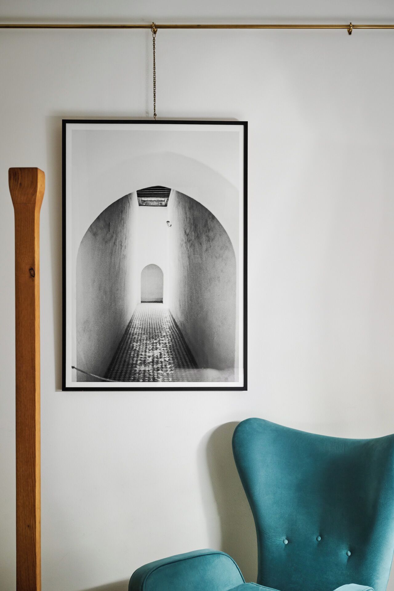 Fotografía en blanco y negro enmarco de un pasillo con techo abovedado y suelo estampado, colgada sobre una pared blanca, encima de un sillón tapizado en color verde azulado.