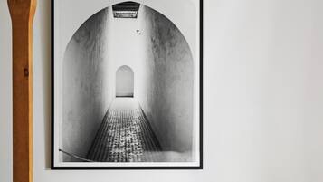 Fotografía en blanco y negro enmarco de un pasillo con techo abovedado y suelo estampado, colgada sobre una pared blanca, encima de un sillón tapizado en color verde azulado.