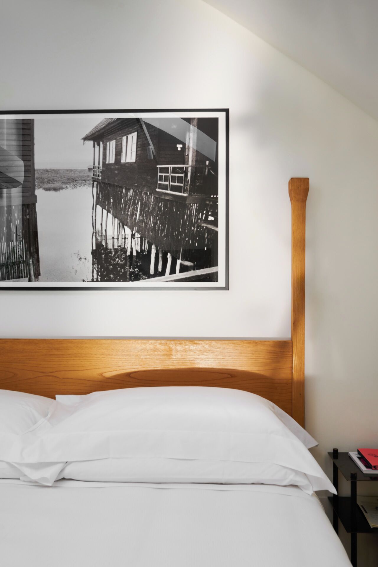 Ein Hotelzimmer mit einem hölzernen Kopfteil, einem Bett mit weißer Bettwäsche und einem schwarzen und weißen Rahmenfoto eines Hauses auf Stelzen über dem Wasser.