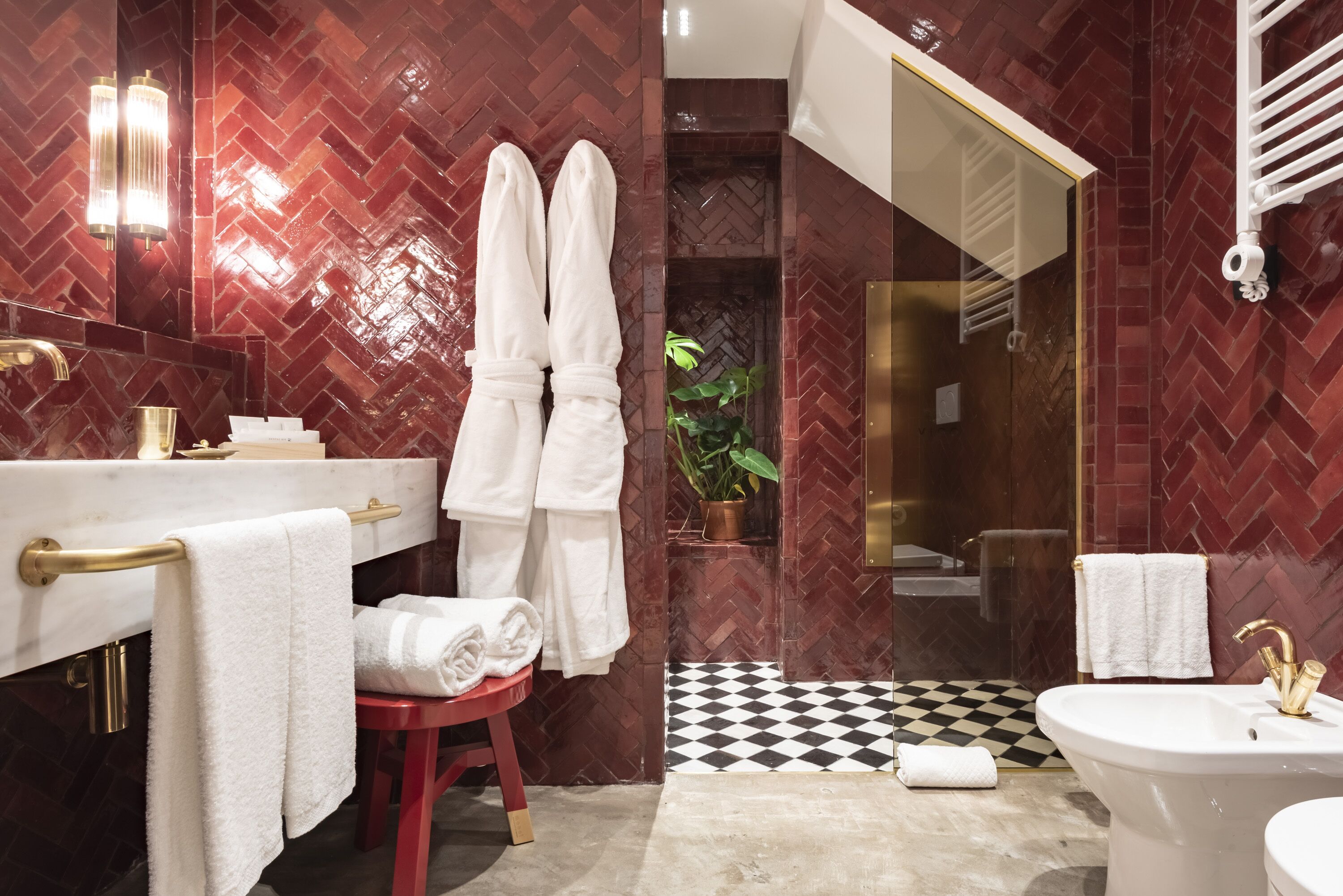 A modern bathroom with a herringbone patterned wall, a gold vanity, a white bathtub, and a red stool.