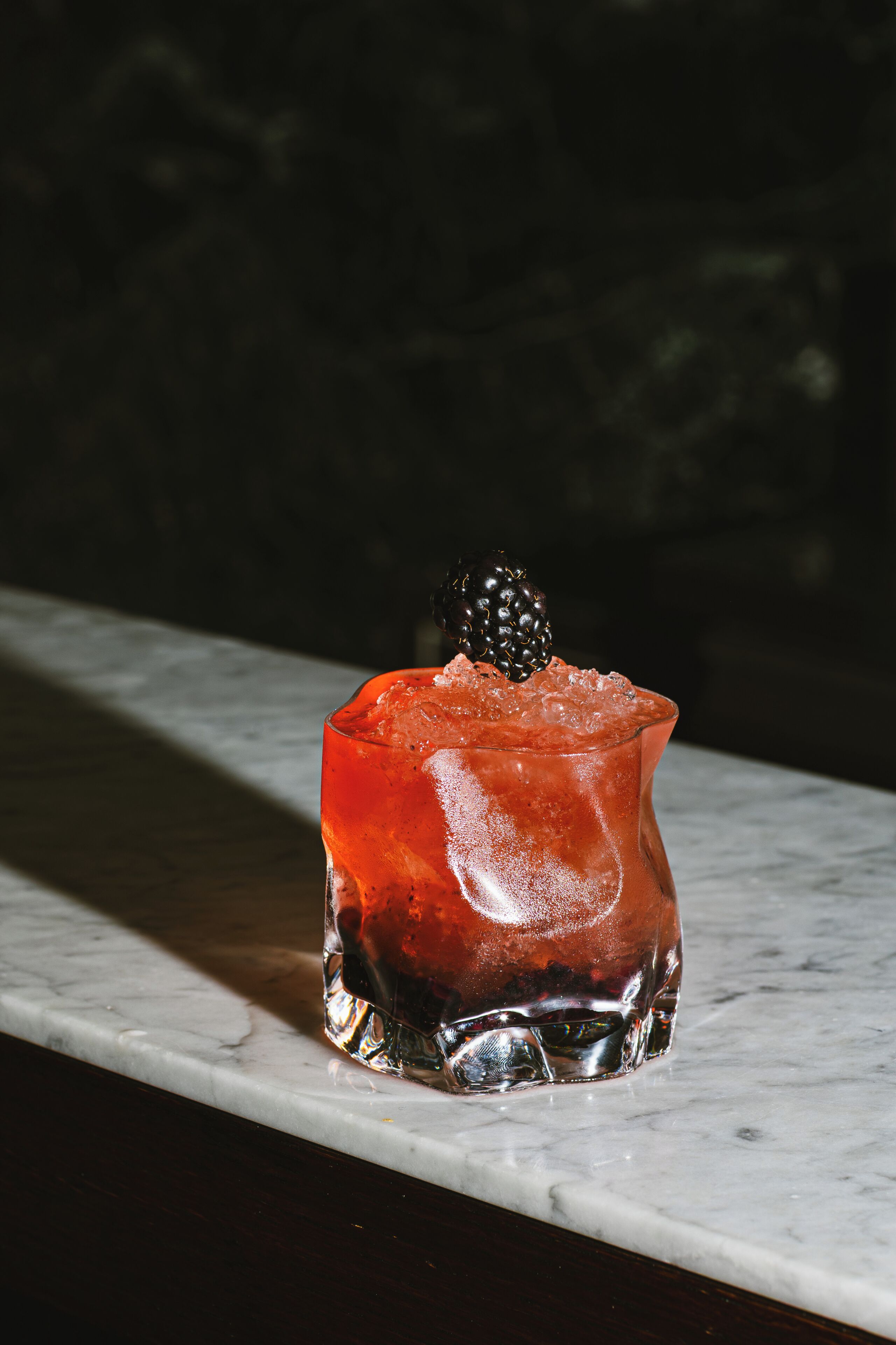 Un verre contenant une boisson rouge, des glaçons et une mûre, posé sur une surface en marbre.