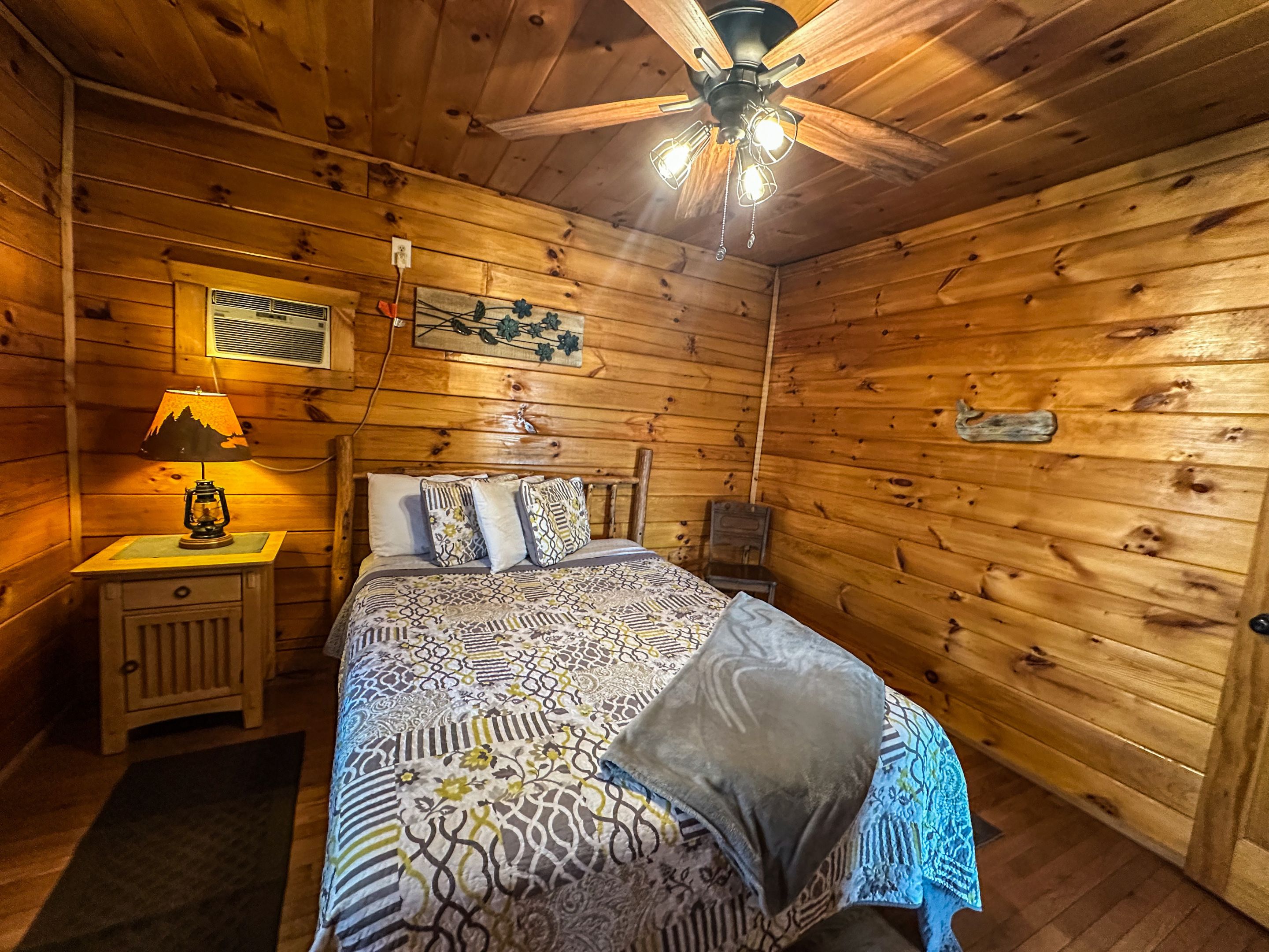 Une chambre dans une cabane en bois, avec un lit, une table de chevet, une lampe et un ventilateur de plafond.