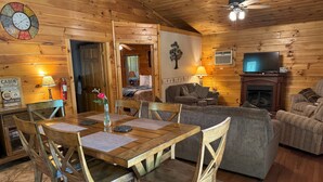 A cabin interior with a wooden dining table, chairs, a sofa, a television, and a fireplace.