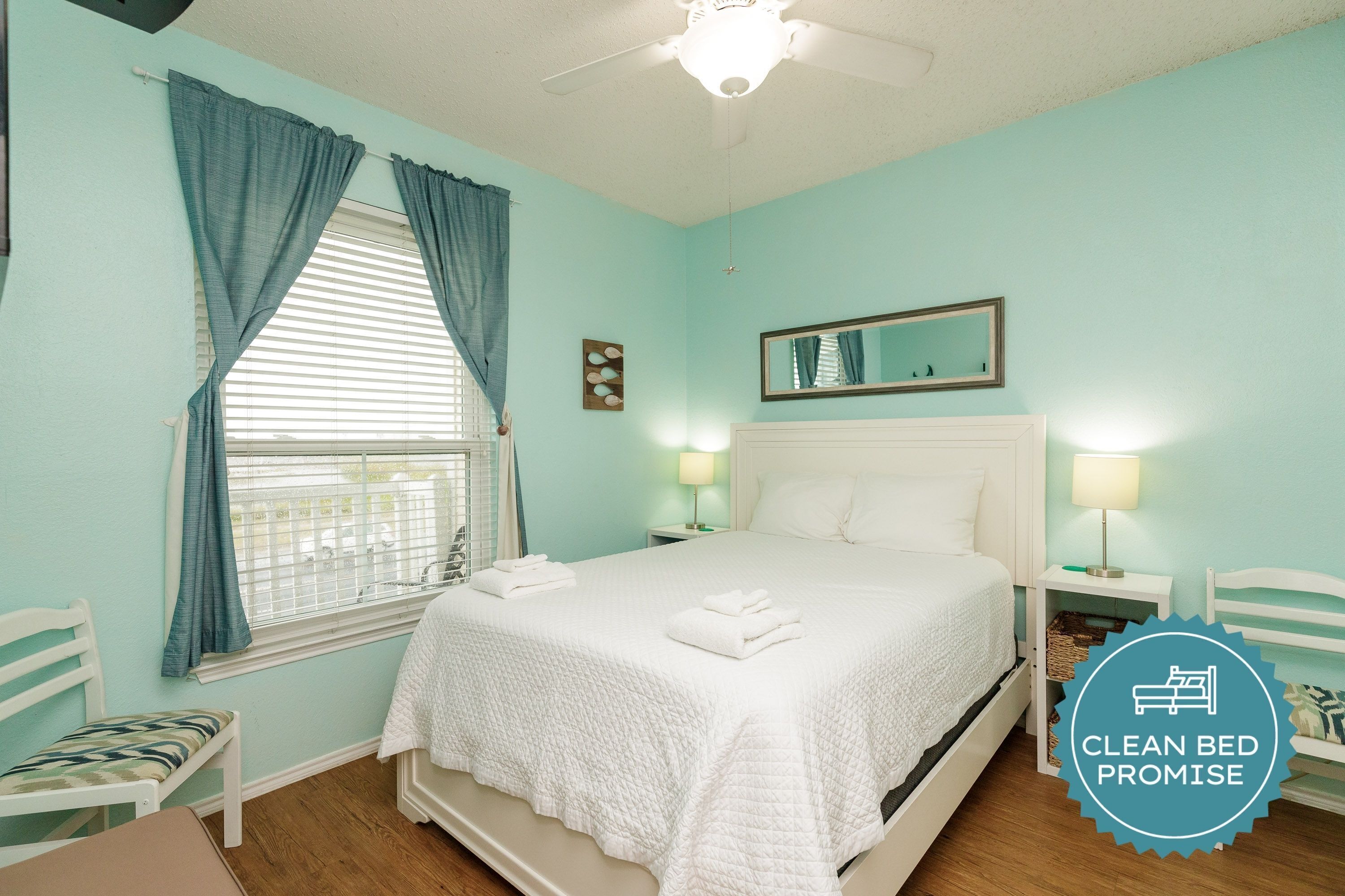 A bedroom with a bed, two bedside tables, a nightstand, a window with curtains, a ceiling fan, and a mirror on the wall.