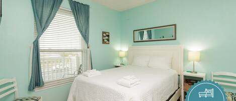 A bedroom with a bed, two bedside tables, a nightstand, a window with curtains, a ceiling fan, and a mirror on the wall.