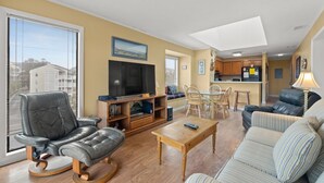 A living room with a leather chair, a television, a dining table with chairs, and a kitchen area in the background.