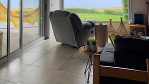 A spacious room with a high ceiling, large windows offering a view of the sea, a comfortable gray chair, a small round table with a plant, and a black pendant light. - Villa Mariense welcomes you in a landscape designed by nature! (Santa Barbara - Santa Maria - Azores Island)