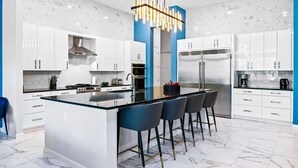 A modern kitchen with white cabinets, a central island with bar stools, a stainless steel refrigerator, and a unique pendant light.