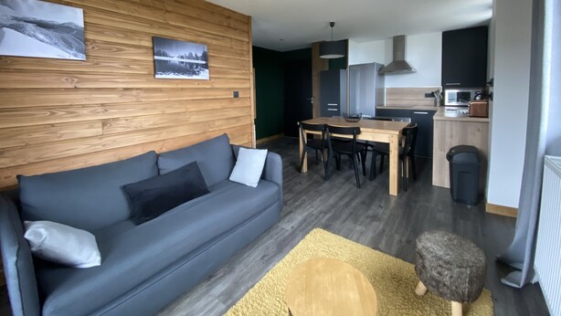 A modern living room with a grey sofa, wooden wall paneling, and a dining area with a wooden table and chairs. - 4-6 Pers apartment in the heart of the Chartreuse at the foot of the ski slopes (Sarcenas)