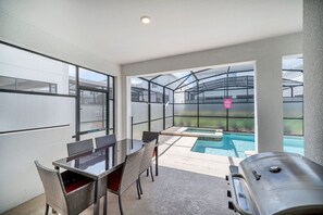 Een terras met een glazen zwembad, een barbecue en een eettafel met stoelen.