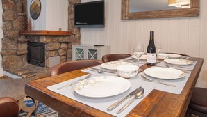 A dining area with a stone fireplace, a flat-screen TV mounted above it, and a wooden dining table set with plates, cutlery, and a bottle of wine.