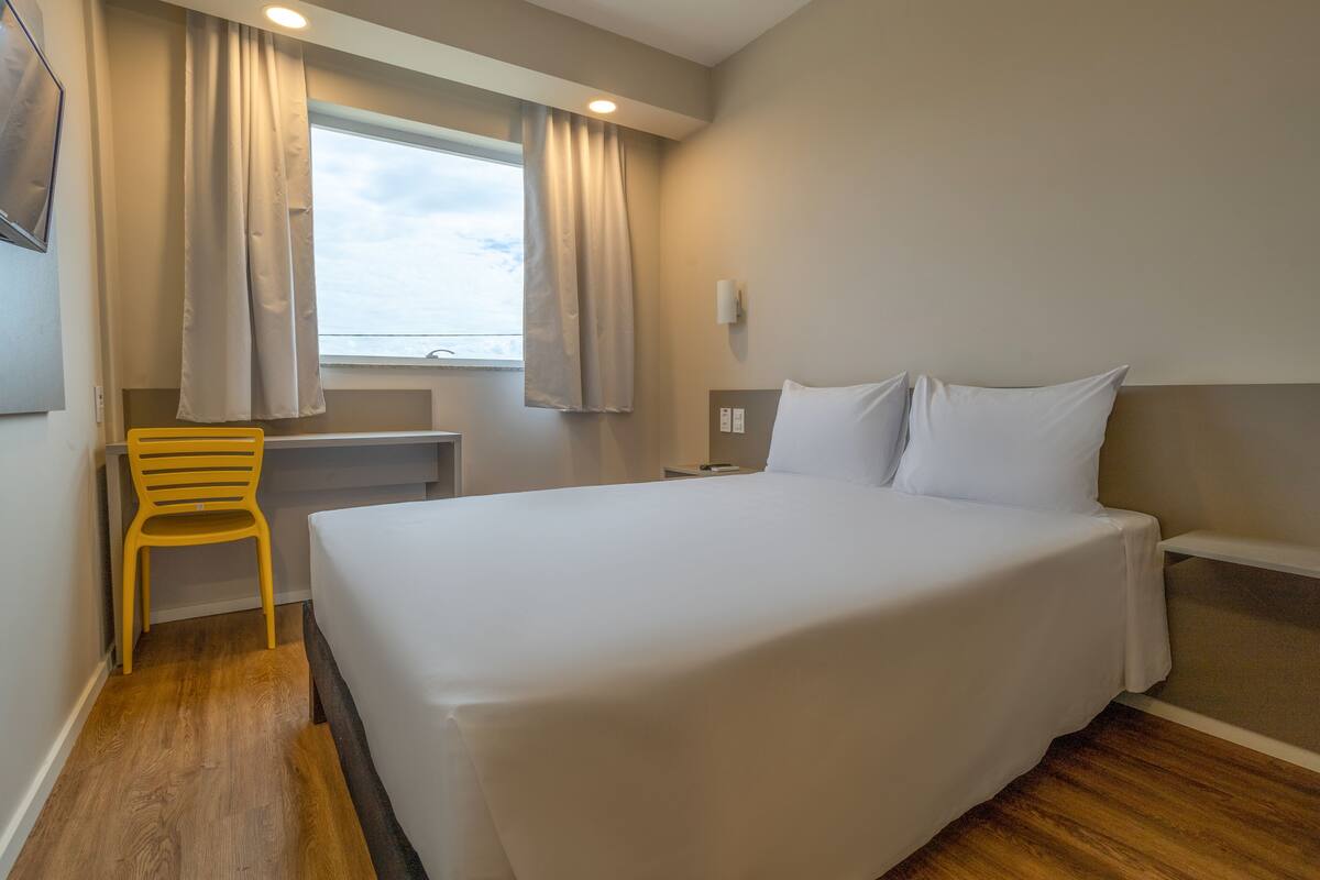 A hotel room with a large bed, a desk, a chair, and a window with curtains.