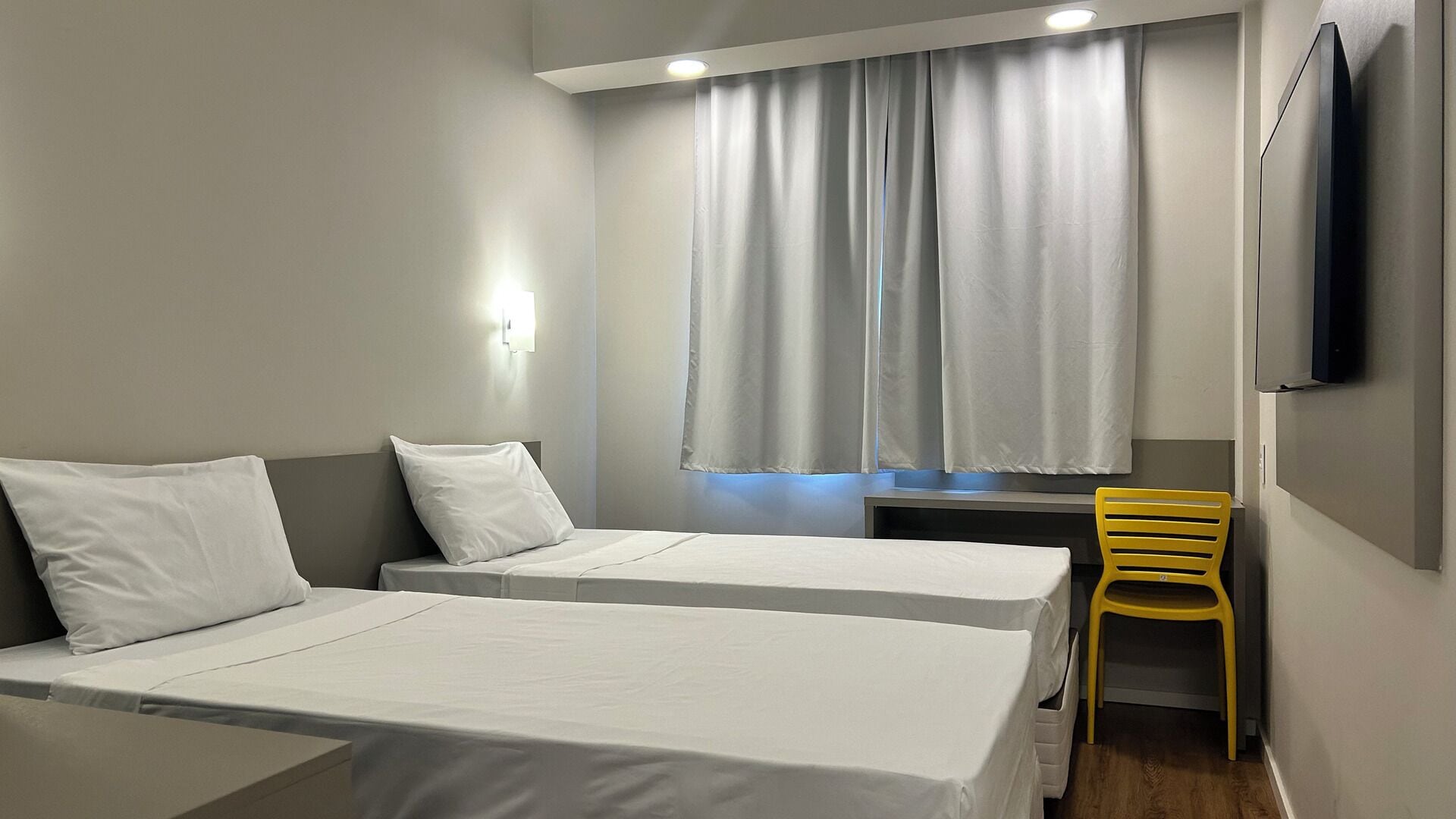 A hotel room with two single beds, a desk, a chair, and a TV mounted on the wall.