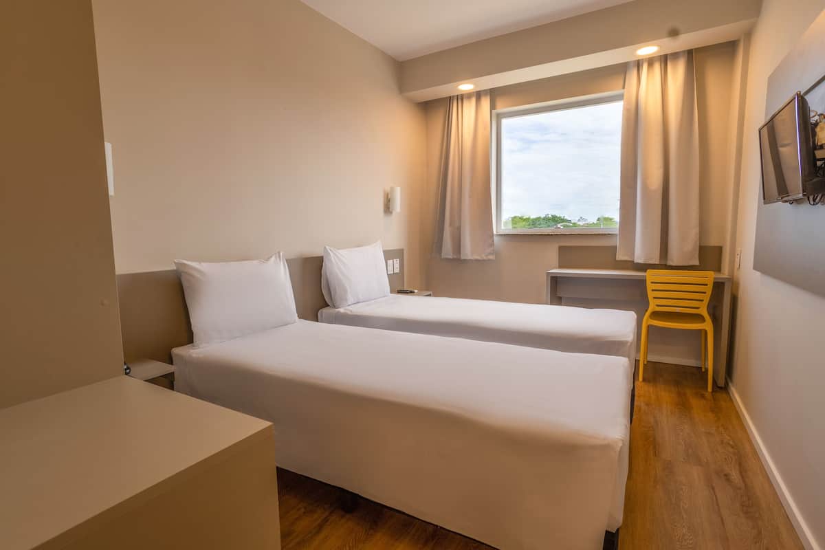 A hotel room with two single beds, a desk, a chair, a TV mounted on the wall, and a window with curtains.