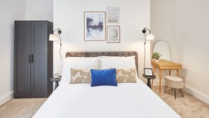 A bedroom with a bed, a nightstand, a desk, a chair, a wardrobe, and wall art.