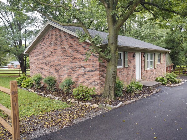 A brick house with a sloped roof, a front porch, and a small garden area with shrubs and a tree. - Ticky's Cottage at Empire Estate (Lebanon)