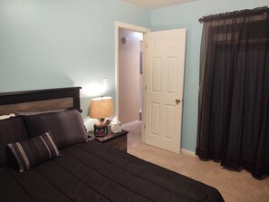 A bedroom with a bed, a nightstand with a lamp, a clock, and a door leading to another room. - Ticky's Cottage at Empire Estate (Lebanon)