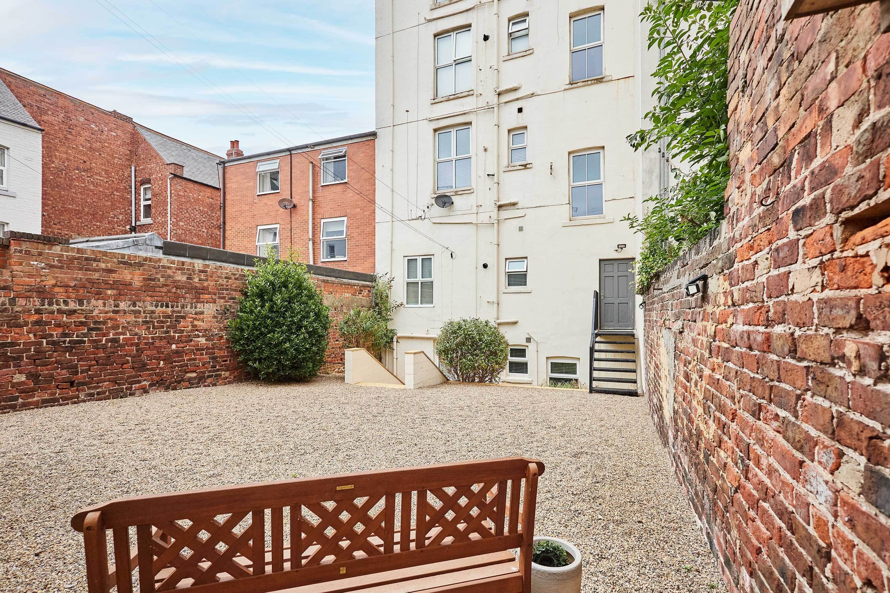 A courtyard with a wooden bench, gravel ground, brick walls, and a multi-story building with windows.