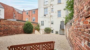 A courtyard with a wooden bench, gravel ground, brick walls, and a multi-story building with windows.