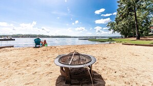 Una playa con un hogar a leña en primer plano, un lago al fondo y una persona sentada en una silla.