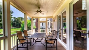 Un patio cubierto con mesa y sillas, ventilador de techo y ventanas amplias que dan a un jardín y edificios.