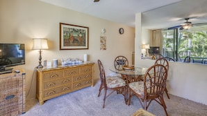 A dining area with a glass table, wicker chairs, a wooden buffet, a television, and a ceiling fan.