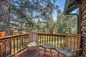 Una terraza de madera con dos sillas y una mesa pequeña, rodeada de árboles y una casa al fondo.