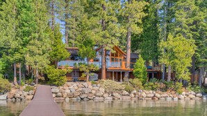 A modern cabin with a wooden deck leading to a lake, surrounded by dense trees.
