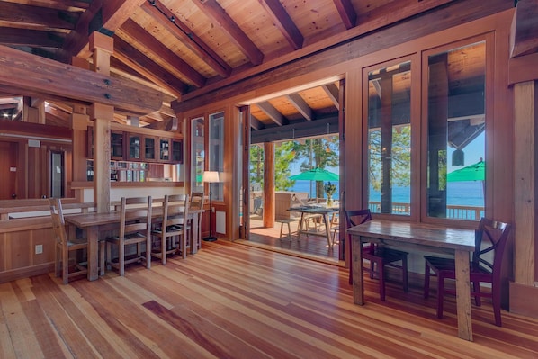 A wooden interior with a dining area, kitchen, and large glass doors leading to an outdoor deck with a view of the sea.
