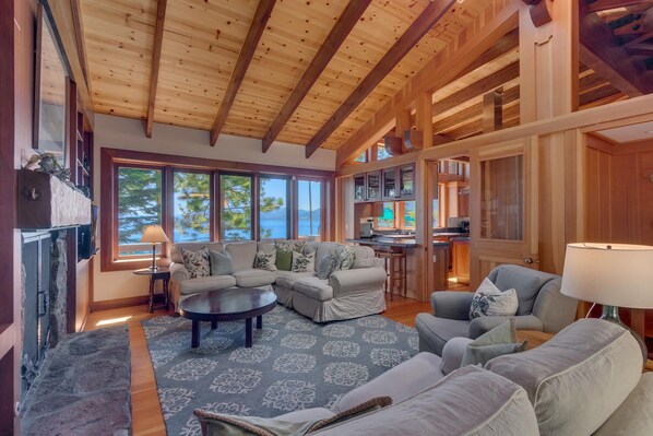 A cozy living room with a fireplace, large windows, and a kitchen area.