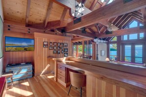 A wooden interior with a bar area, framed pictures on the wall, and a large window overlooking a body of water.