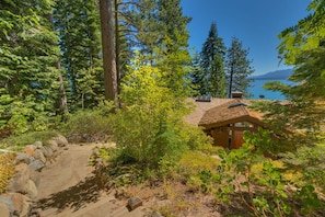 A cabin with a sloped roof, surrounded by dense forest and a clear view of the lake.