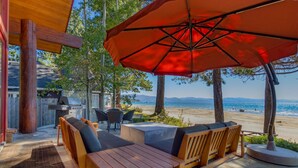 Een terras buiten met houten meubels en een rode parasol, met uitzicht op een strand en bergen.