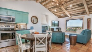 Una cocina con gabinetes verde azulado, una mesa de comedor con sillas, una araña y una vista por la ventana.