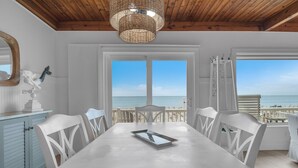 A dining area with a view of the beach, white chairs, a long table, and a wicker chandelier.