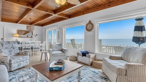 A spacious living room with a wooden ceiling, large windows overlooking the ocean, and a cozy seating area with a coffee table.