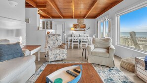 A spacious living area with a high wooden ceiling, a dining area with white furniture, and a large window offering a view of the sea.