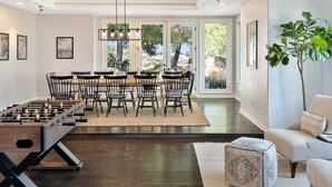 A dining area with a foosball table, a large dining table with chairs, and a view of the outdoors through large windows. - Vineyard View by Fieldtrip 8 Bd with Infinity Pool, Hot Tub, Games (Murrieta)