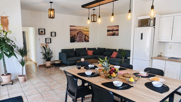 A modern living room with a wooden dining table set for a meal, a sofa, and a kitchen area with appliances. - Spacious T5, terrace, near center, private parking (Perpignan)
