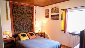 A bedroom with a large bed, two bedside tables with lamps, a tapestry on the wall, and a window with curtains. - Casa do Olival - Double room with exterior toilet-Indian (Barreira Grande)