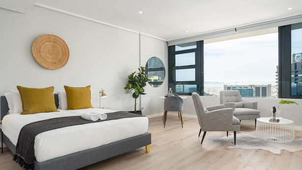 Une chambre d’hôtel moderne avec un grand lit, deux fauteuils, une petite table et une vue sur la ville.